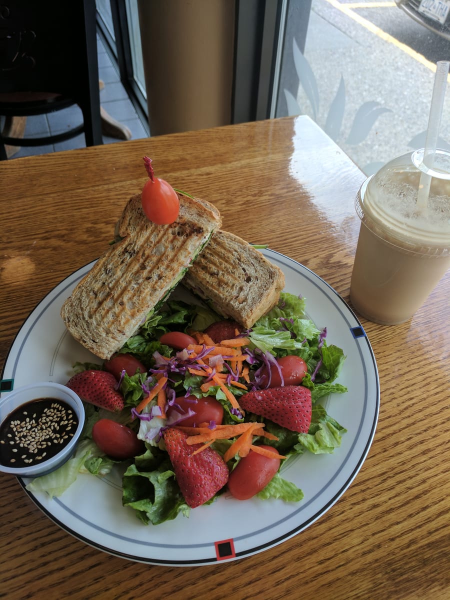 A grilled sandwich cut in half with a cherry tomato pick, plated with green salad, strawberries and shredded carrot, beside an iced drink on a wooden table