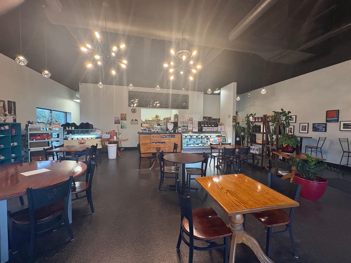The dining room at Starberry Cafe: wooden tables and dark chairs under pendant and sputnik lights, with the service counter, display case and chalkboard menu at the back