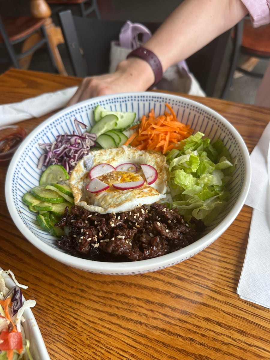 A rice bowl with seasoned beef, a fried egg topped with radish slices, lettuce, carrot, cucumber and red cabbage, on a wooden table