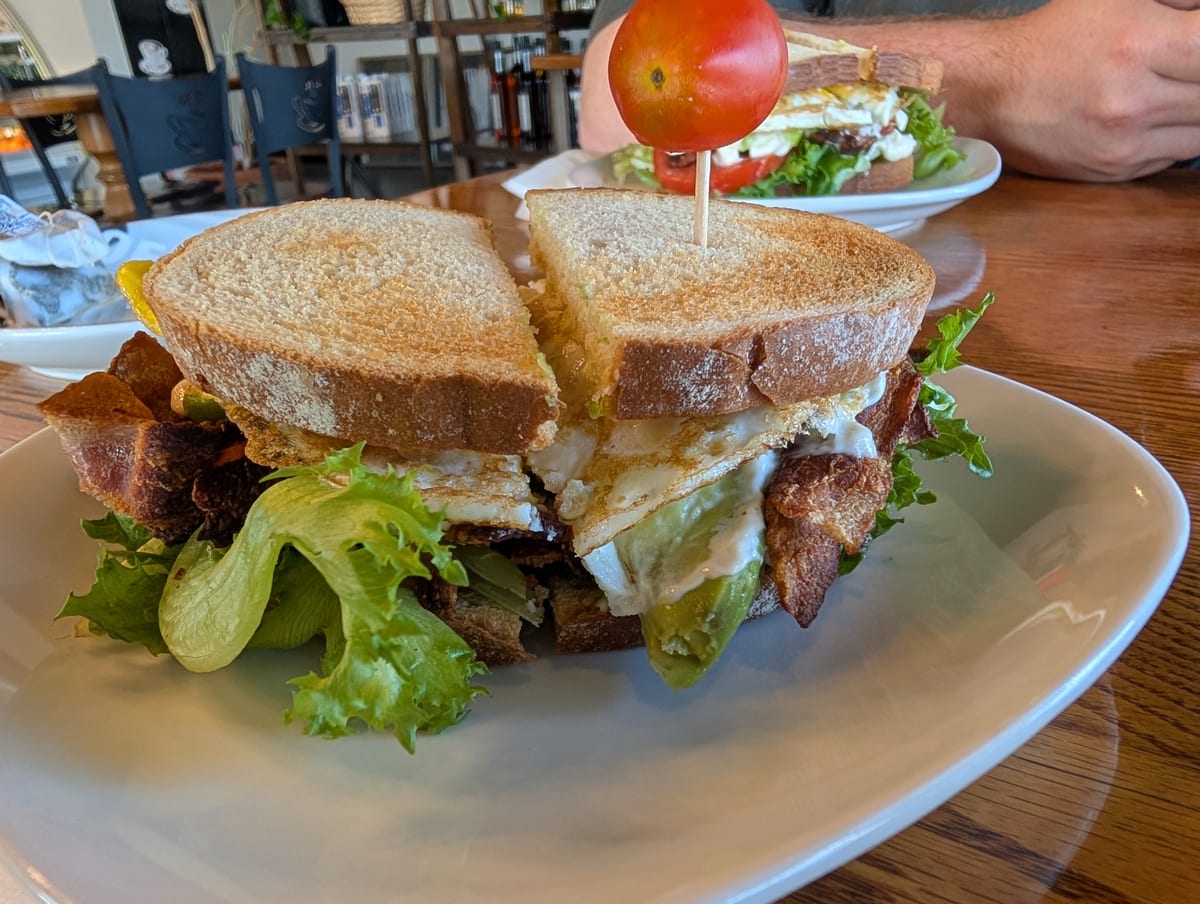 Close-up of a halved sandwich with bacon, fried egg, avocado and lettuce on toasted bread, a tomato on a pick beside it